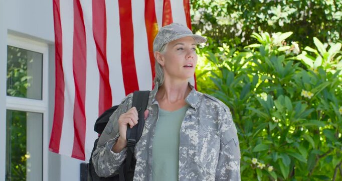 Camera pulling in, woman in camo holding backpack strap, US flag on porch, scanning before leaving