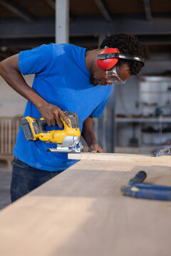 Carpenter is leaning on workshop bench using yellow jigsaw cutting wooden plank under bright lights