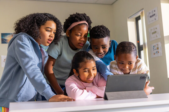 Five diverse children leaning over tablet in gray folio case on white table in classroom