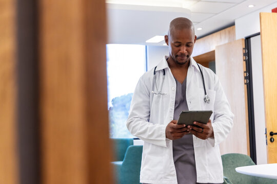 African American adult male doctor standing in clinic wearing lab coat and stethoscope using tablet