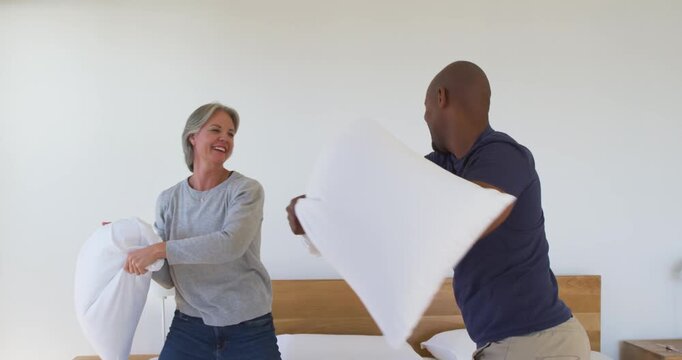 Diverse couple picking up white pillows for fun, swinging, blocking, tumbling on bed with headboard