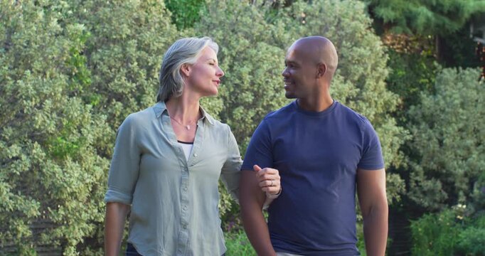 Diverse couple linking arms walking in garden, woman speaking, couple talking together, green shirt