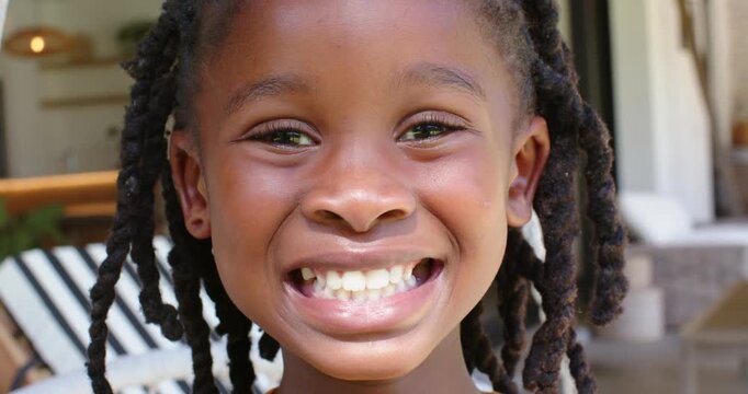 African child smiling while camera gaze widening grin on porch swing, braids bobbing, mustard top