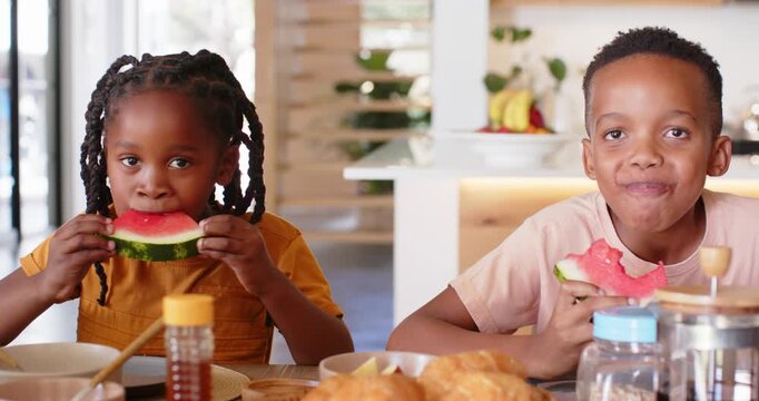 Sibling children seeing croissants and taking watermelon slices for breakfast at table in kitchen