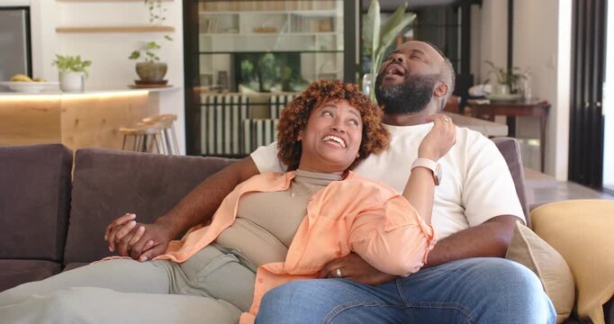 African couple holding hands on U-shaped sofa, woman laughing, leading man nuzzling temple