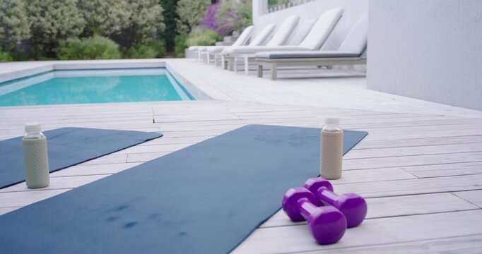 Camera is moving forward and right, centering mat with bottle and dumbbells for workout by pool