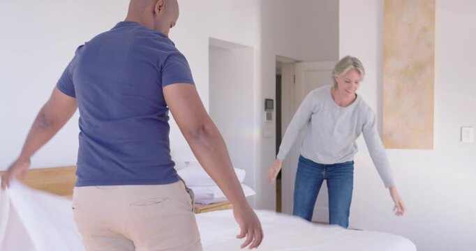 Diverse couple lifting white sheet, shaking, draping bed, smoothing corners while making bed