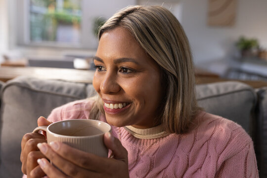 African American woman in pink sweater holding ceramic mug smiling on living room sofa
