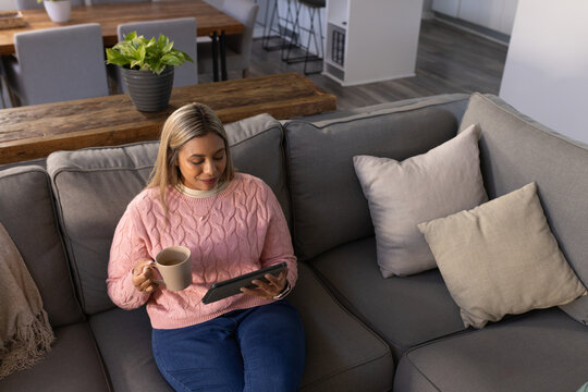 Mid adult African American woman sitting on gray sofa holding tablet and mug, in pink sweater