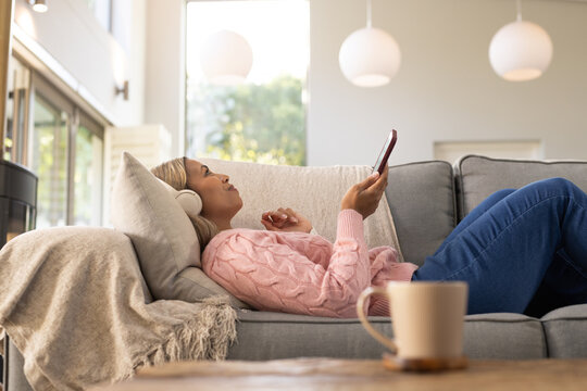 Middle-aged African woman reclining on sofa at home wearing pink sweater holding phone with headset