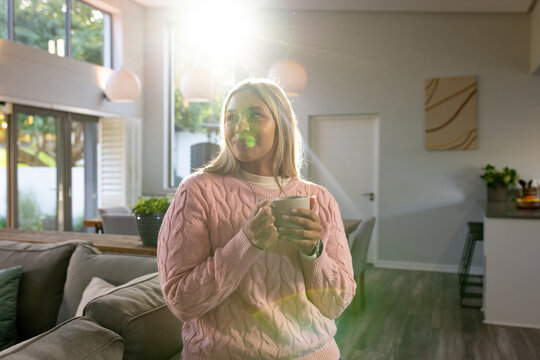 African American middle-aged woman holding ceramic mug, wearing pink sweater in sunlit living room