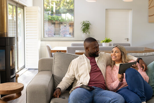 African American couple sitting closely on light gray sofa holding tablet and remote in living room