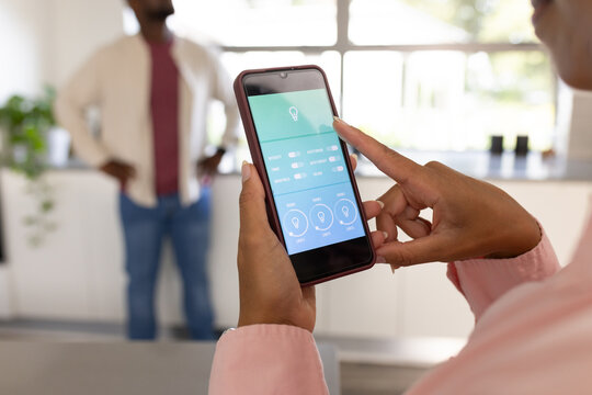 African American couple using smartphone, controlling lighting app in bright kitchen with plant