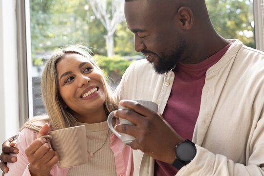 Diverse couple leaning at home by large window holding ceramic mugs, wearing pendant and wristwatch