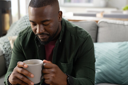 Adult African American man sitting on gray sofa at home holding ribbed mug, wearing green shirt