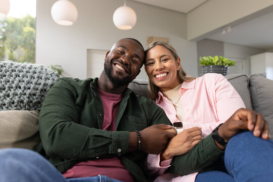 Diverse couple sitting closely on grey sofa in living room, showing smartwatch and bracelet