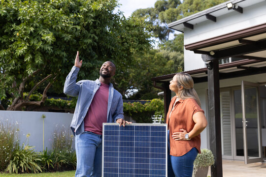 African American couple standing in backyard by solar panel, pointing upward and smiling