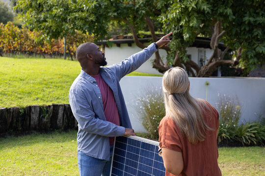 Mid-adult man and woman holding large solar panel, pointing toward roof on residential lawn
