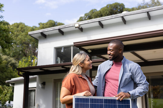Diverse couple holding solar panel while smiling and chatting on covered patio at modern house