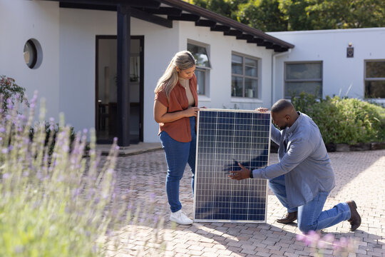 African American woman holding solar panel in rust shirt while man tech kneeling on brick driveway