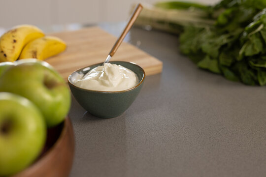 Small green bowl holding thick cream, spoon resting on gray counter, green apples, bananas blurred