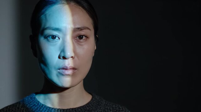 Asian woman face illuminated by dual split lighting. Half of face lit by blue light while other half is in shadow. Concept of duality and contrast in portrait