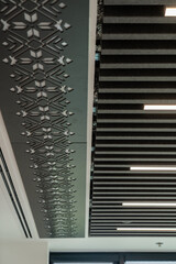 Fototapeta premium A modern office ceiling features a perforated geometric pattern beside dark acoustic baffles with integrated linear lights, blending functional sound control and minimalist style for a contemporary co