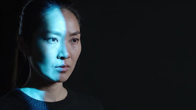 Asian woman illuminated by blue light on her face. Serious female expression with dramatic split lighting. Cinematic facial portrait for technology and concept