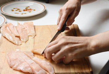 Obraz premium Close-up of hands cutting raw chicken fillets in a modern kitchen on a wooden cutting board. Background shows plates and cooking ingredients, creating a home cooking and food preparation concept.