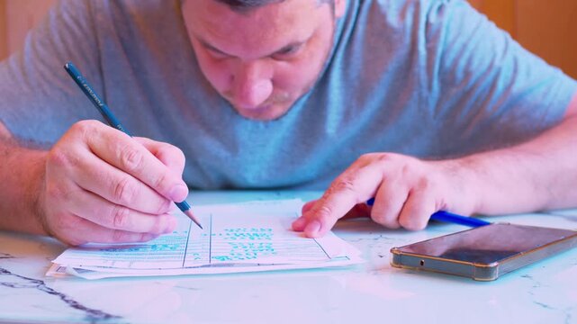 Low angle close-up of a focused man filling out a paper document.Home office.Cinematic shot of a middle-aged man concentratedly writing on a form or contract at a desk under natural light.