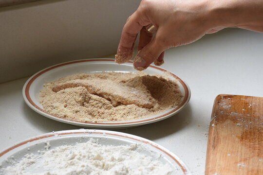 Close-up of a hand breading a chicken fillet in breadcrumbs during food preparation. Plates with flour and coating ingredients are visible on the kitchen workspace. Concept of homemade cooking, meal p
