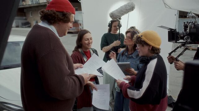 Young female director holding script pages and discussing details with actors before movie shoot. Makeup artist, camera operator and sound technician preparing for next take on outdoor film set