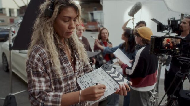 Female production assistant filling out slate clapperboard on outdoor movie set. Director, camera operator, makeup artist and actors preparing for scene in background