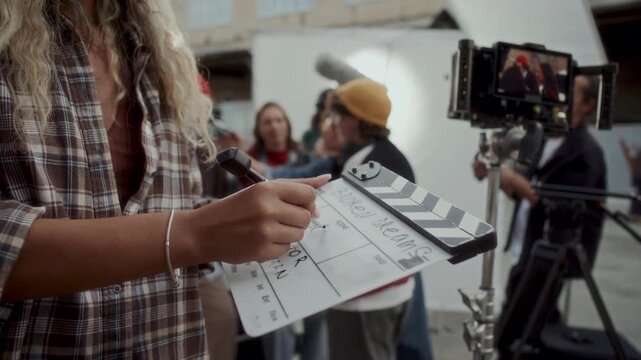 Close up view of hands of female production assistant writing scene details with marker on clapperboard, movie crew members preparing for shoot at outdoor location in background