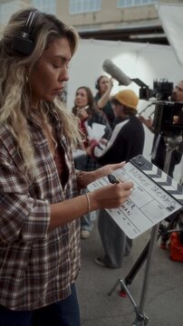 Female film crew member in headset writing production information on clapperboard while preparing for shooting scene on outdoor movie set. Vertical format video