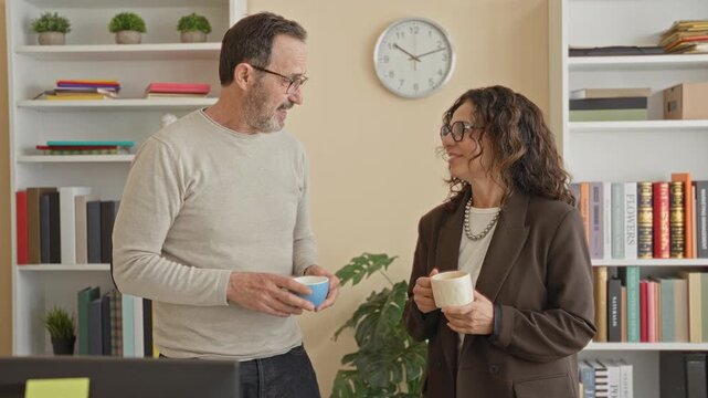 Man holding blue mug and gesturing with finger while talking to woman near bookshelf and wall clock in office; friendly chat.