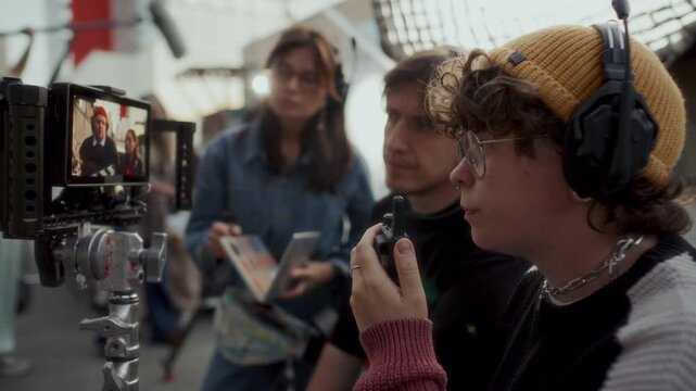 Young female filmmaker talking on walkie talkie and reviewing footage on camera monitor with her crew during independent movie production shoot at outdoor location