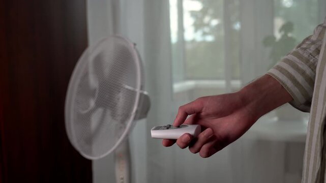 The hand of a young man in a linen shirt turns on an electric floor fan with a remote control during abnormal heat