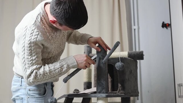 One young handsome Caucasian man installs hanging toys into the hole in the tube with his hands, assembling a new cat house, standing in the living room, side view close-up with slow motion. Concept o