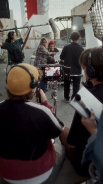 Film production crew watching camera monitor on outdoor set, clapper loader marking scene and boom operator holding microphone above actors during video shoot. Vertical clip