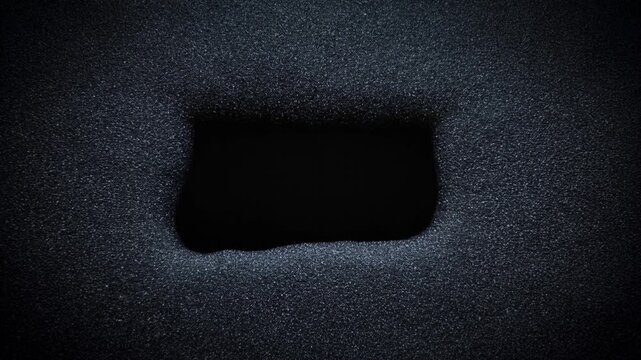 Tracking shot of rectangular void sliding laterally across dark textured granular surface with subtle pan in close top studio view