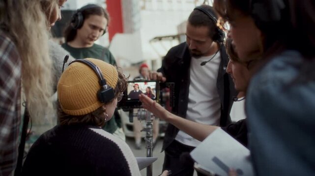 Female film director and her creative production crew discussing footage on camera playback screen during outdoor commercial video shoot
