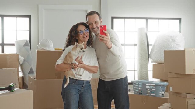Middle aged couple with small dog, man holds red smartphone taking a selfie inside a bright livingroom filled with moving boxes, laundry basket and rolled rug in a building; home joy.