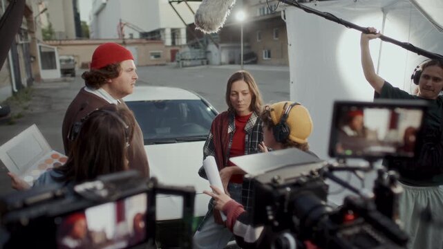 Film director explaining script and assistant slating clapperboard before actors performing emotional drama scene in front of camera on outdoor movie set