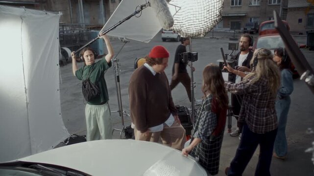 Video production crew working together on outdoor movie set to film scene with two actors, cinematographer operating camera on tripod, boom operator holding microphone, director explaining script