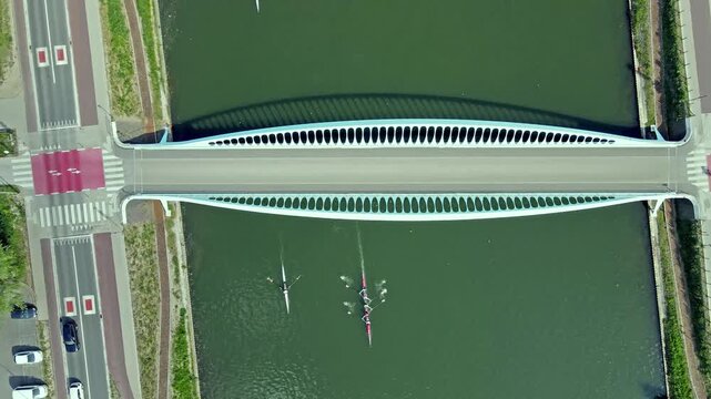 Aerial top-down drone view of rowers passing under a white arched bridge over a green canal with roads and cars on both banks a summer view of urban infrastructure and water sport.
