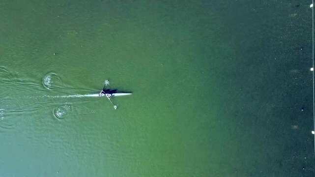 Aerial top-down drone view of a single scull rower gliding over teal green water in summer the boat and oars leaving soft ripples and a thin wake in a minimalist serene water sport scene.