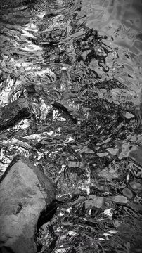 Black and white vertical format of shallow water rippling around big and small rocks, creating circles in a calm atmosphere. 