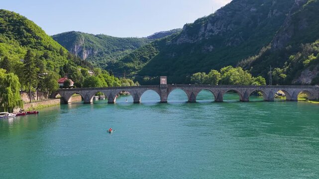 Wide drone shot of Mehmed Pasha Sokolovic Bridge with kayakers on the Drina River and mountains beyond