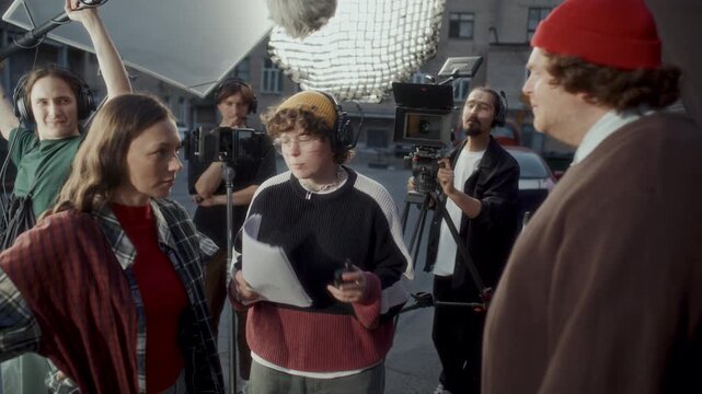 Young actor and actress acting out emotional argument scene during film shoot, professional media production crew surrounding them with cameras, boom microphone and lighting equipment. Zoom in shot